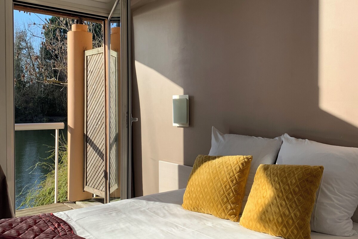 A hotel room with a bed, a window with a view of trees and water, an air conditioning unit, and a wall-mounted light.
