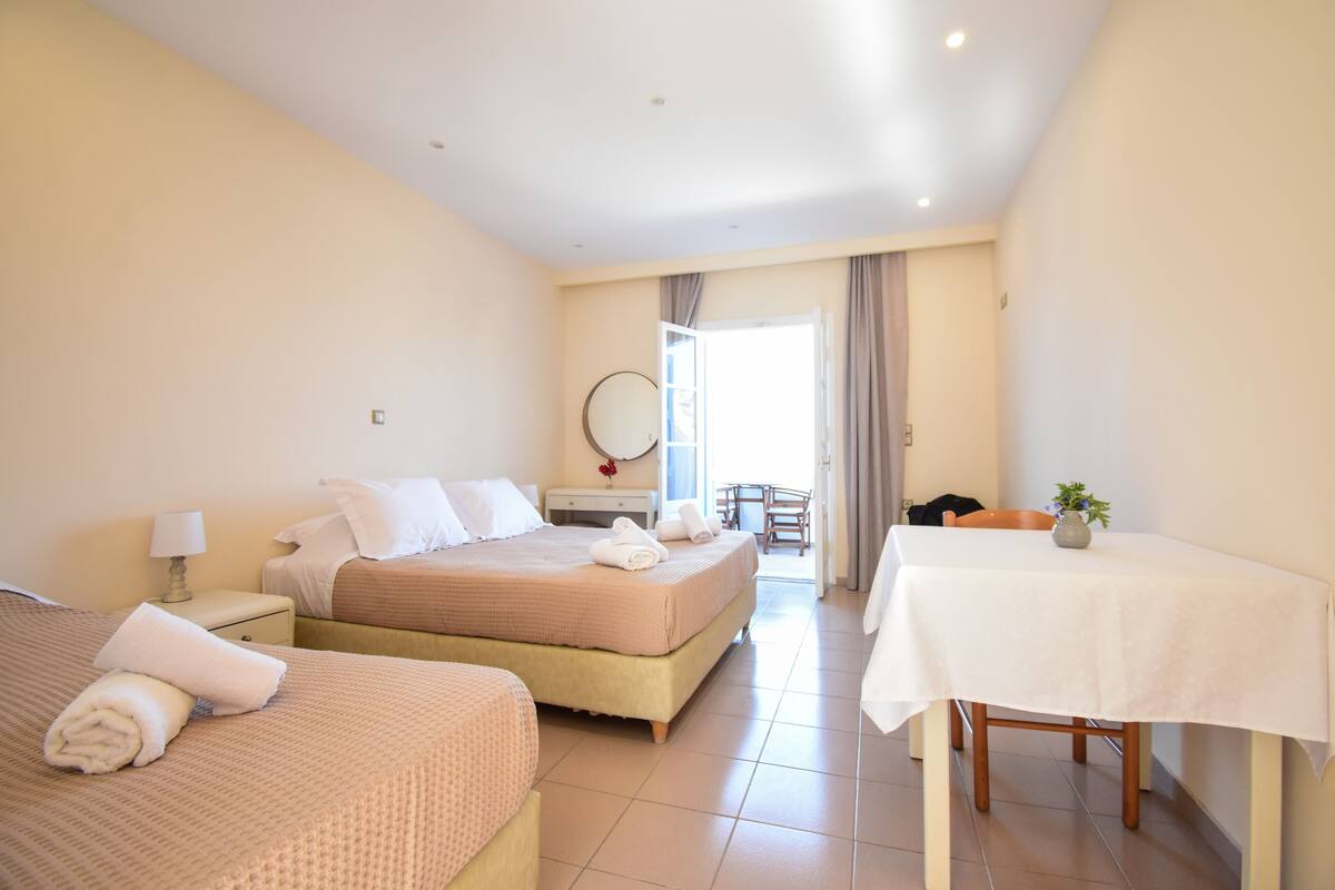 A hotel room with two beds, a dining table, a chair, a small table with a vase, and a view of the outside.