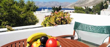 A table with a plate of fruit and a chair with a view of a mountainous landscape and the sea.