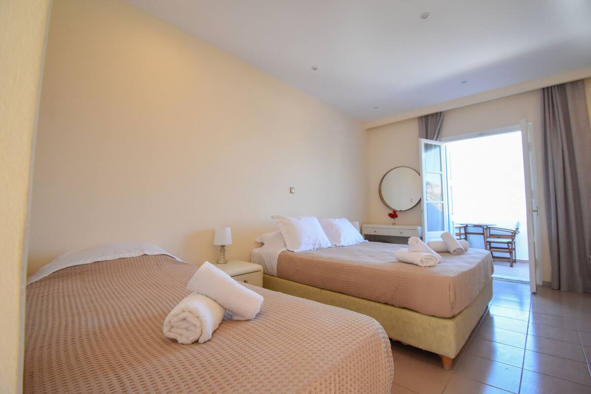 A hotel room with two beds, a small table, a chair, and a balcony with a view.
