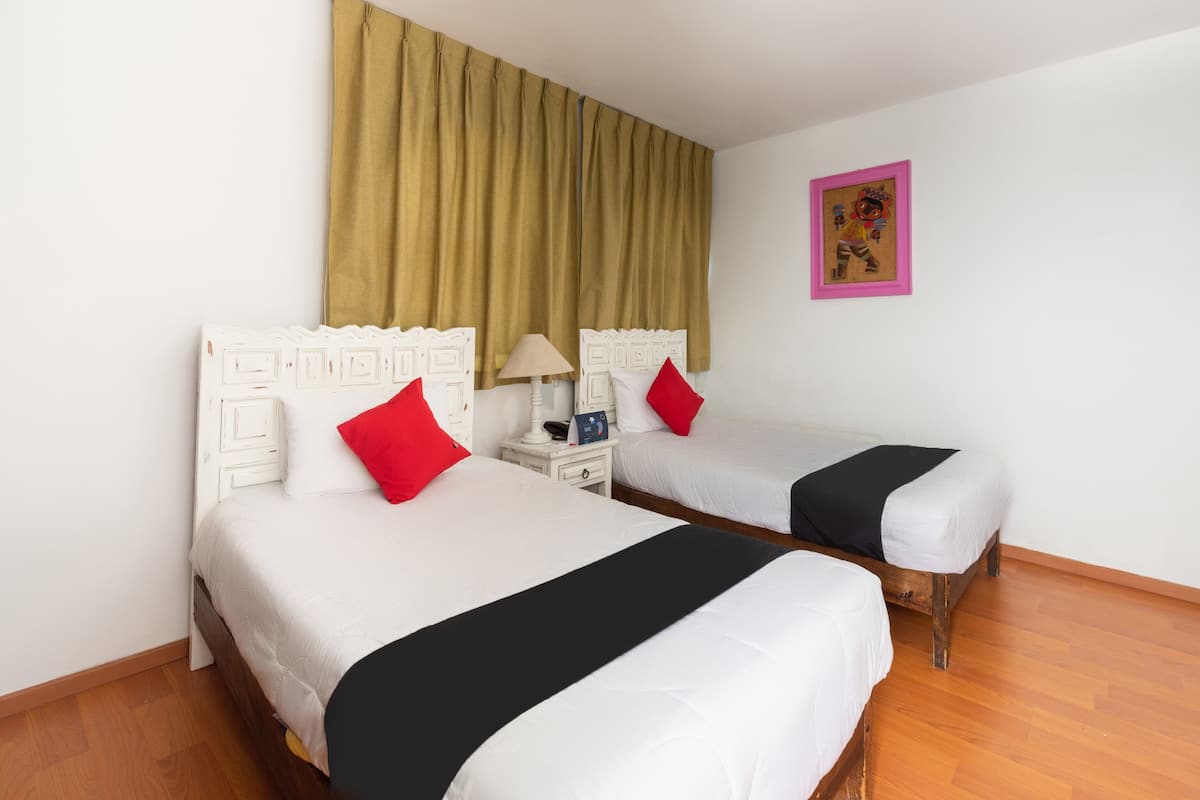 Habitación de hotel con dos camas, una mesita de noche con lámpara, un cuadro en la pared y suelo de madera.