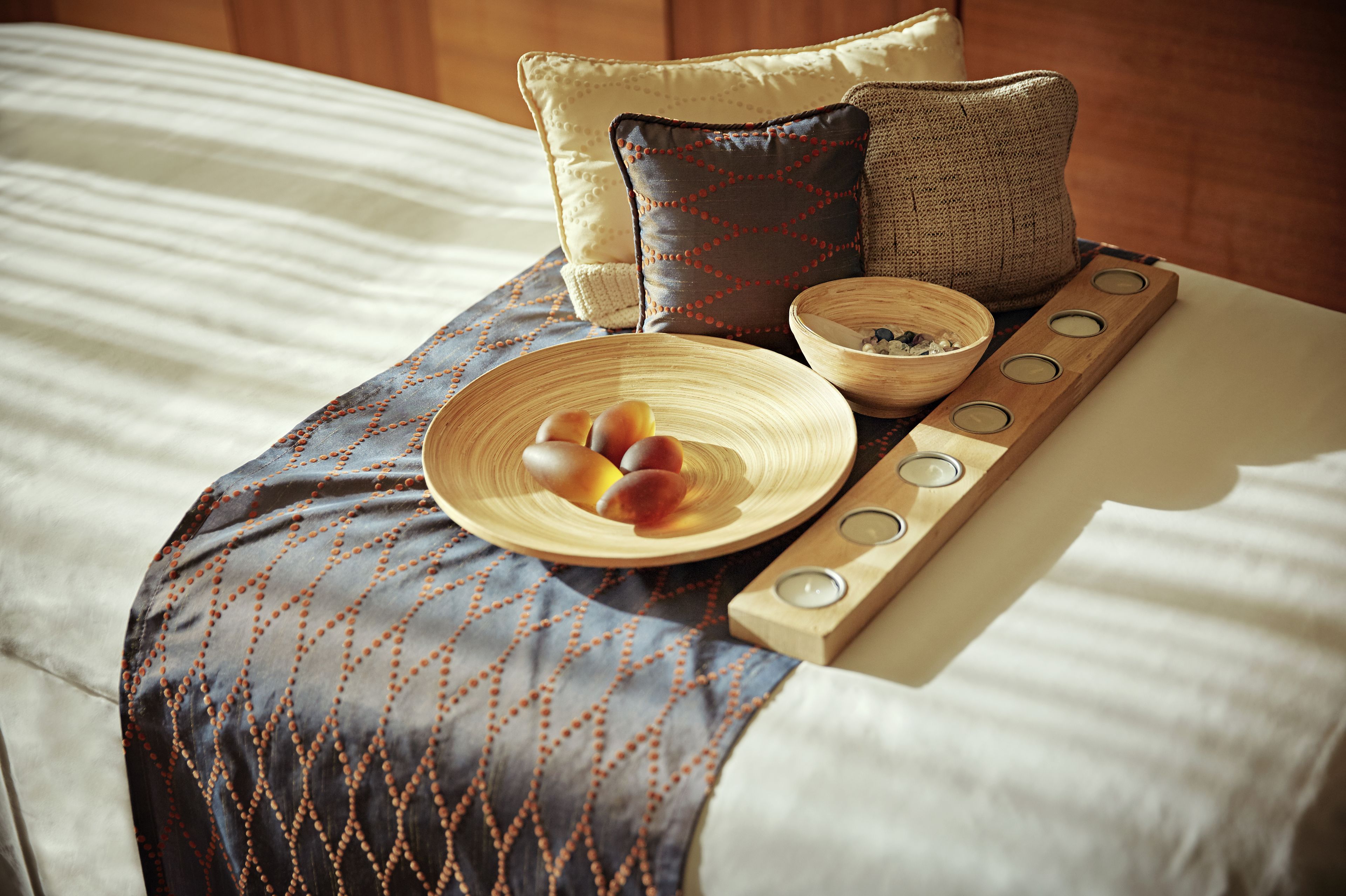 A neatly made bed with a patterned blanket, a wooden tray with candles, and a bowl of fruit.