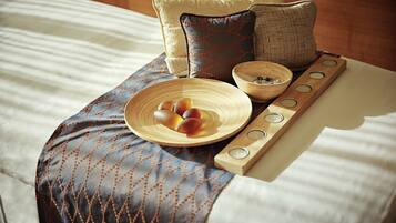 A neatly made bed with a patterned blanket, a wooden tray with candles, and a bowl of fruit.