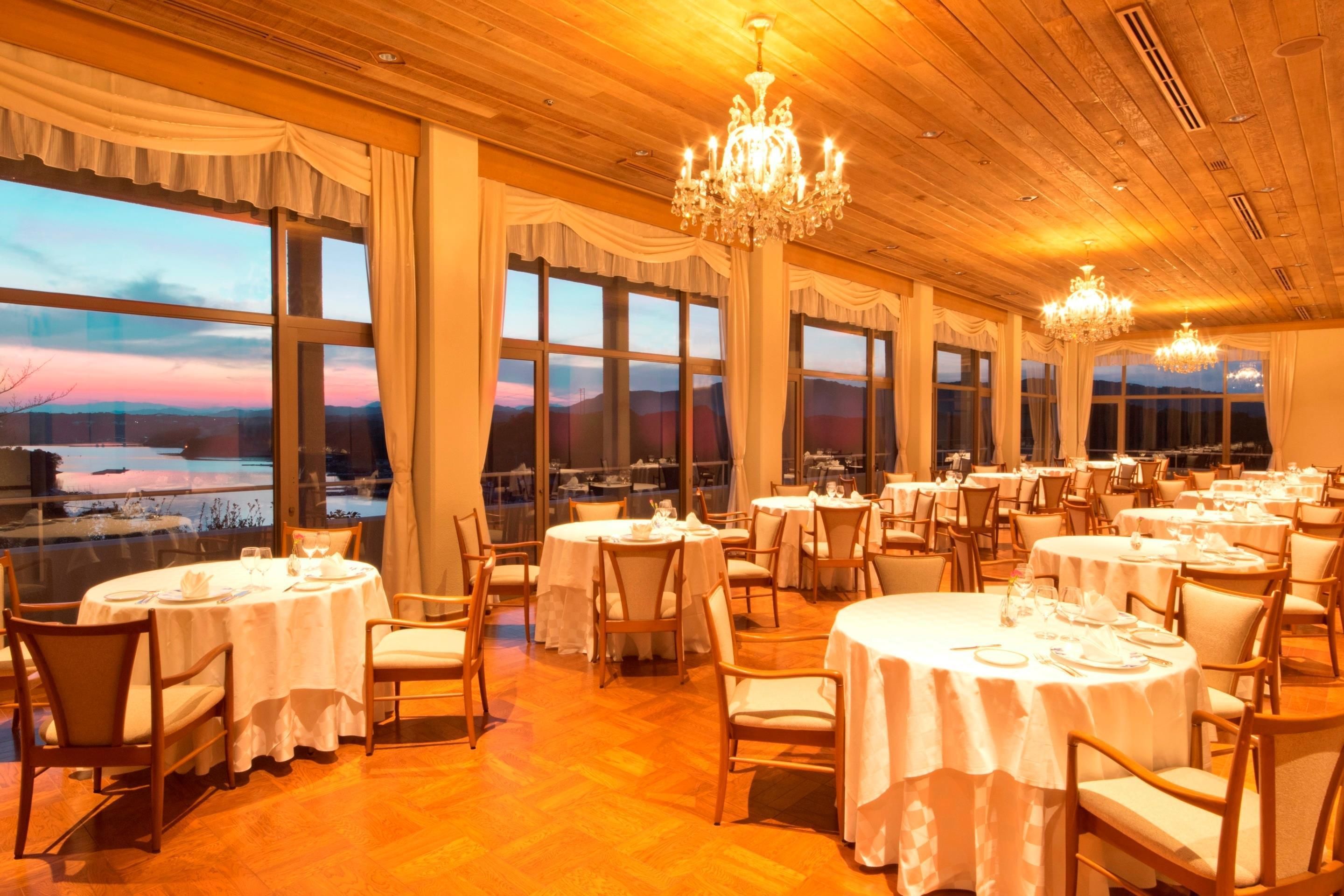 Un restaurante con mesas y sillas redondas, ventanas amplias y candelabros.