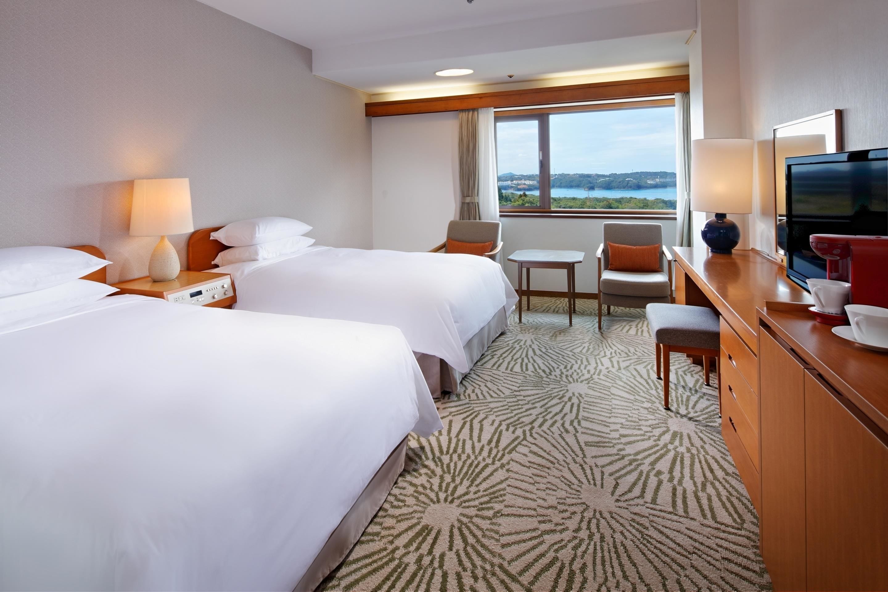 A hotel room with two beds, a small table, a TV, and a view of the outdoors.