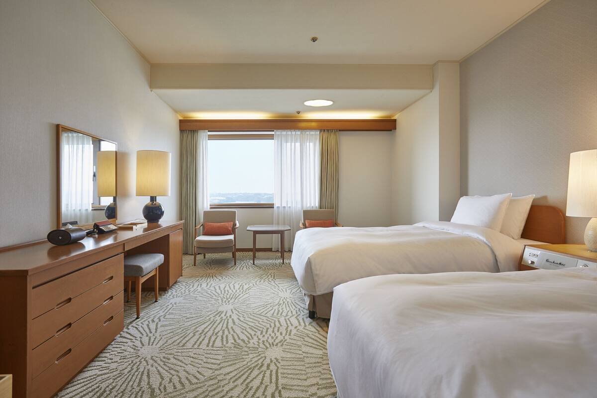 A hotel room with two beds, a desk with a lamp, a chair, a small table, and a view of the outdoors through the window.