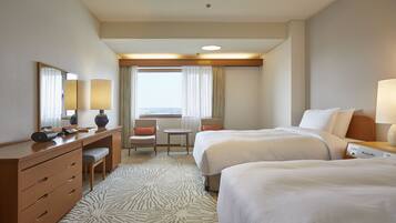 A hotel room with two beds, a desk with a lamp, a chair, a small table, and a view of the outdoors through the window.