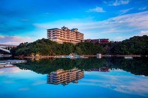 Un hotel de varios pisos con balcones, ubicado en una colina con vista a una masa de agua tranquila.
