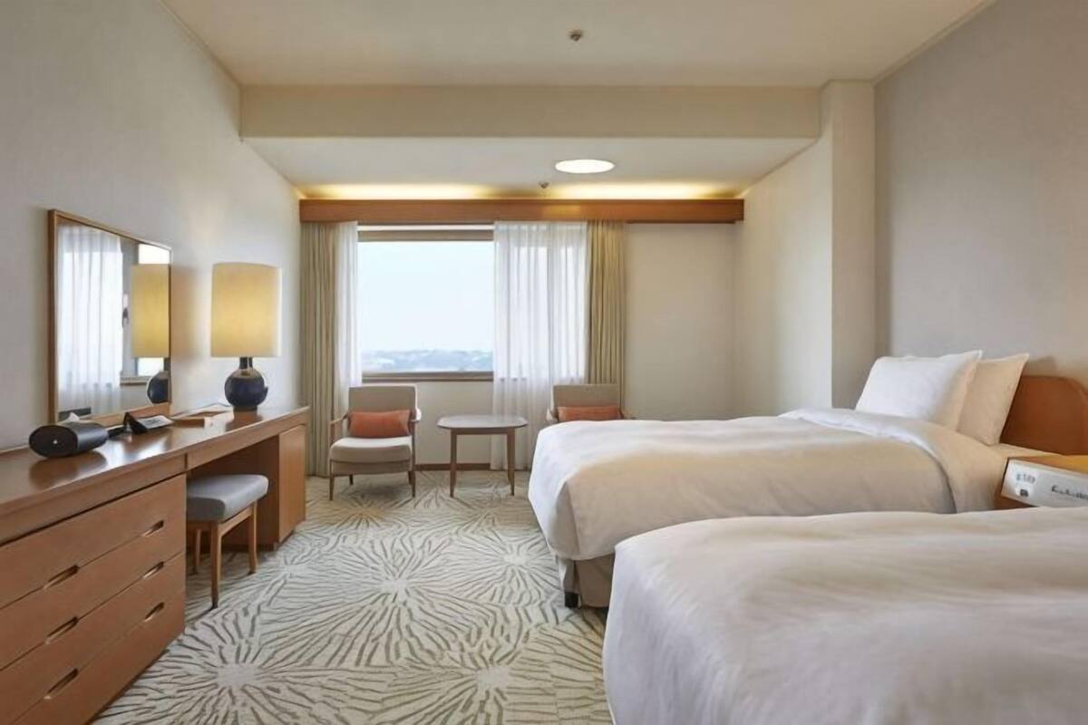 A hotel room with two beds, a desk with a chair, a small table, and a window with curtains.