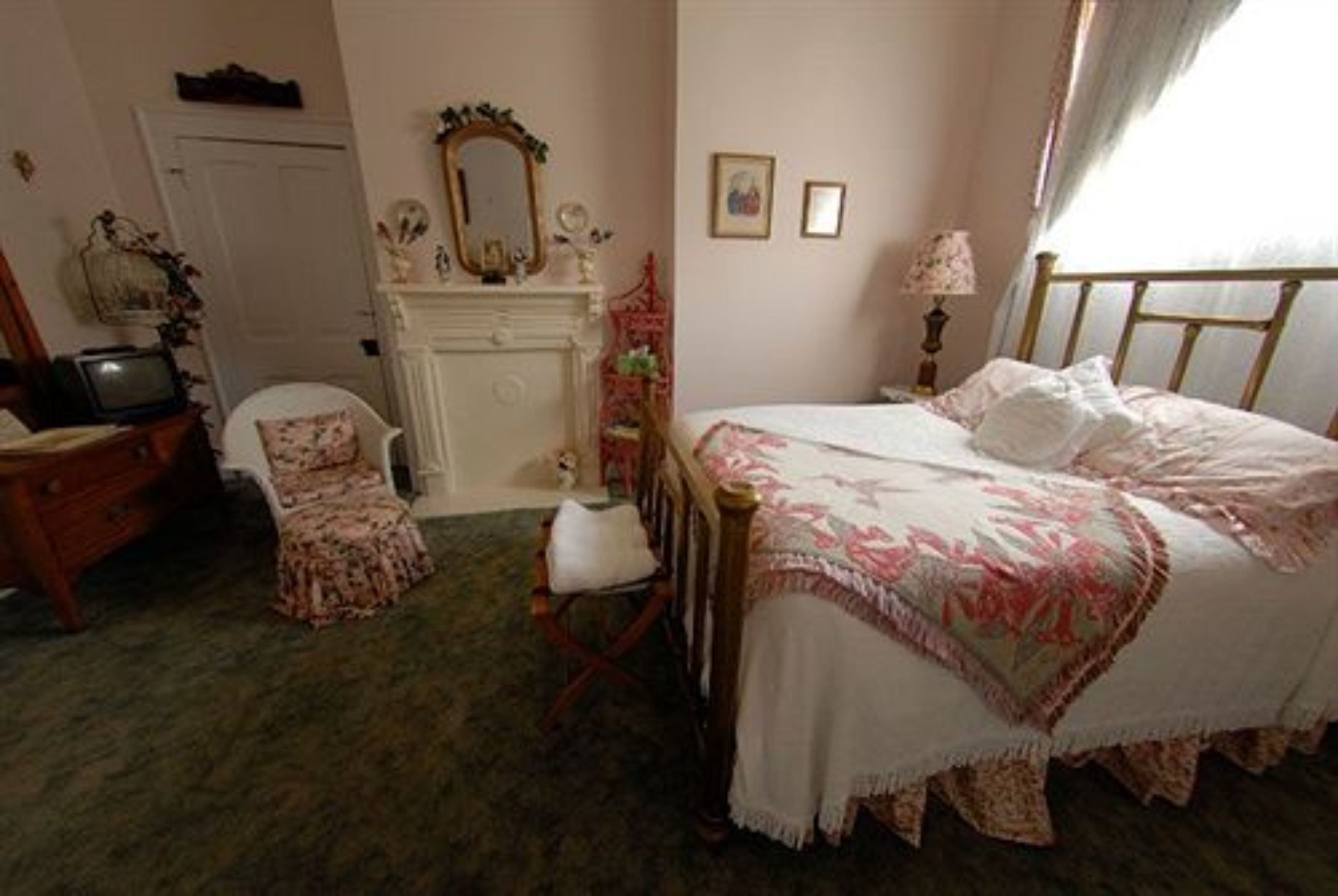 A bedroom with a bed, a dresser, a chair, a vanity with a mirror, and a fireplace.