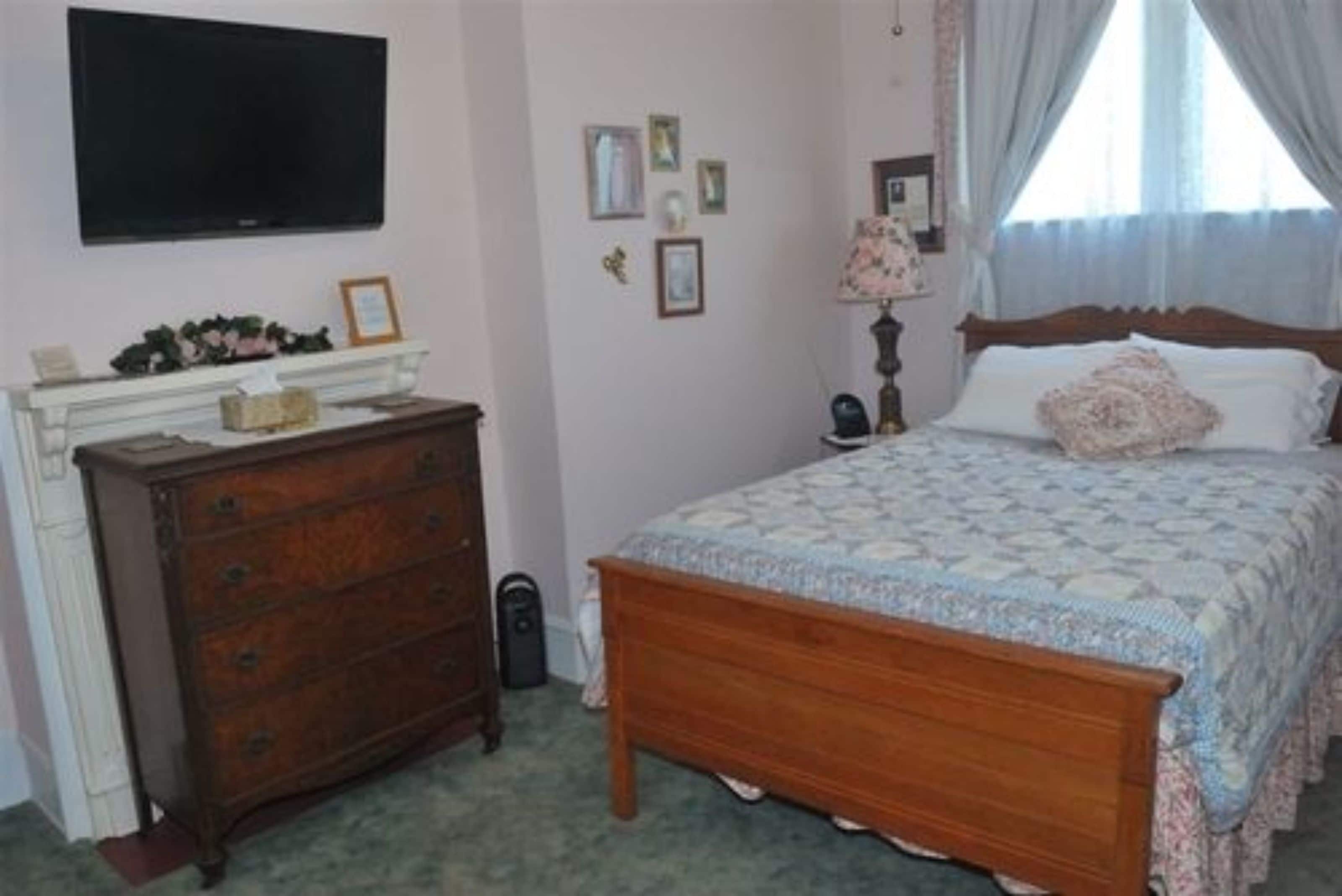 A bedroom with a wooden bed, a dresser, a television mounted on the wall, and a window with curtains.