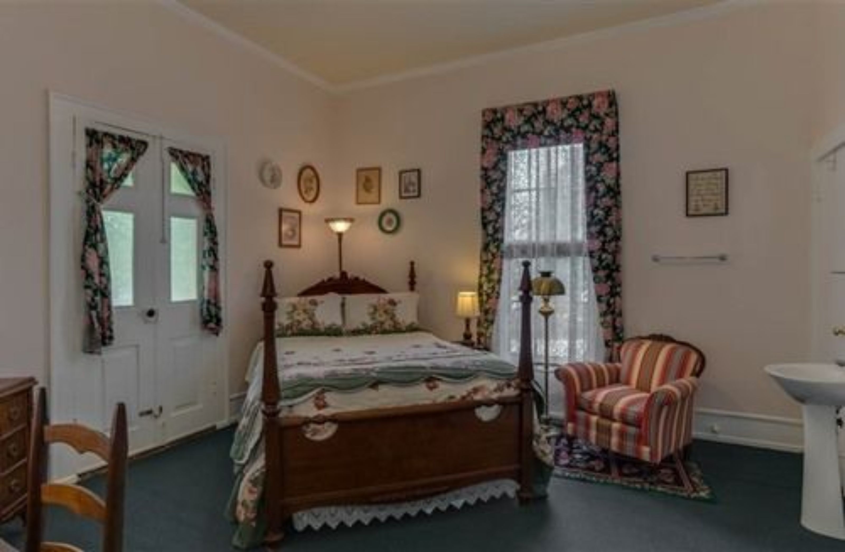 Ein Schlafzimmer mit einem hölzernen Bett, einem Sessel, einem Nachttisch, einer Lampe und einem Fenster mit floralen Vorhängen.