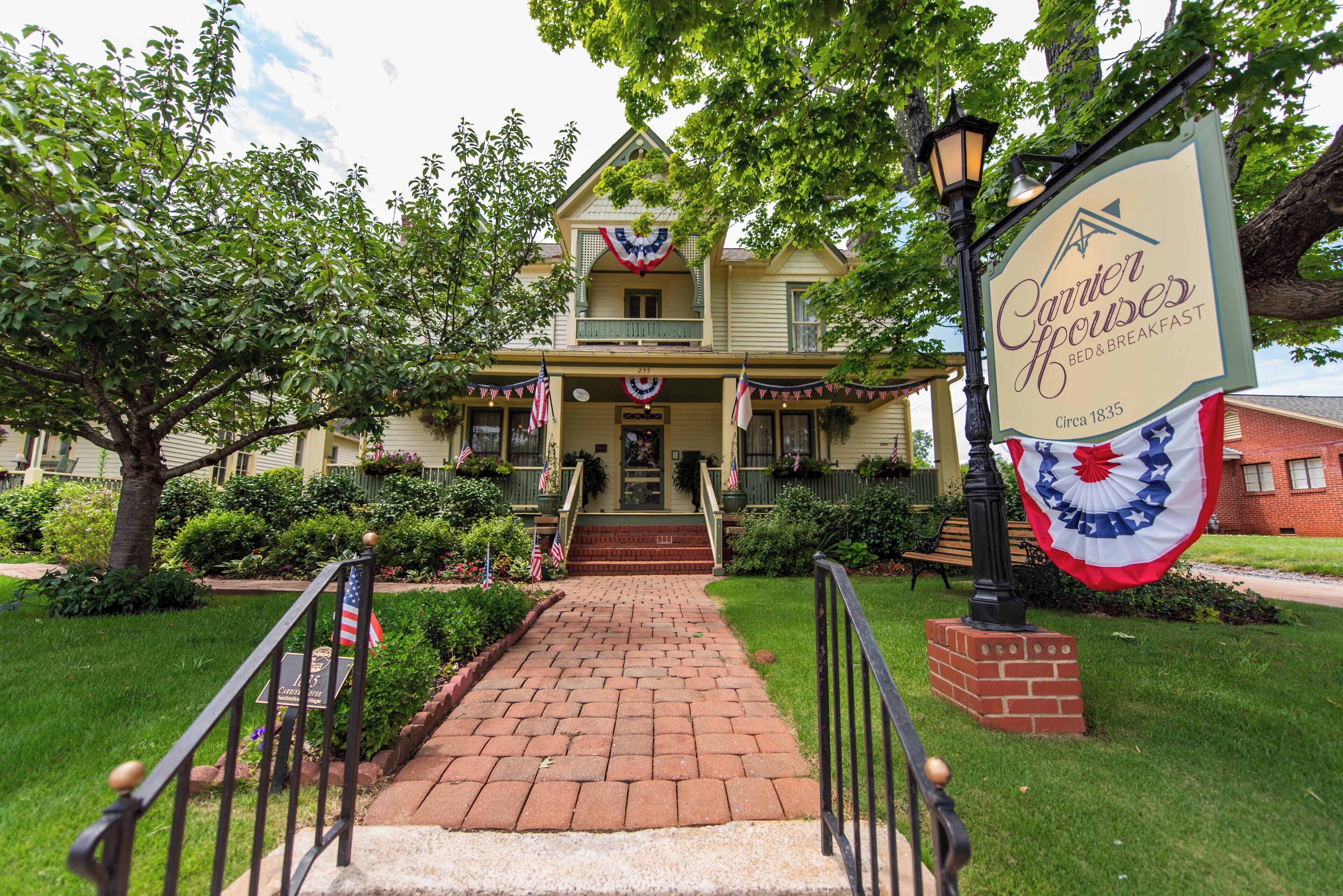 A historic bed and breakfast with a brick pathway leading to the entrance, surrounded by greenery and American flags.