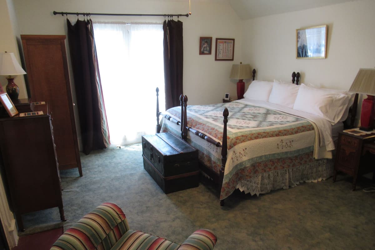 A bedroom with a large bed, a wooden dresser, a chair, and a window with curtains.
