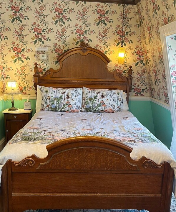 A wooden bed with floral bedding, a nightstand with a lamp, and floral wallpaper. - Angel of the Sea (Cape May)