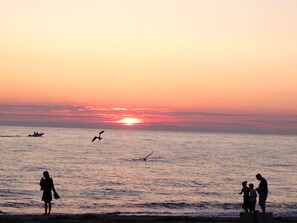 A sunset over a calm sea with silhouettes of people and a boat. - Angel of the Sea (Cape May)