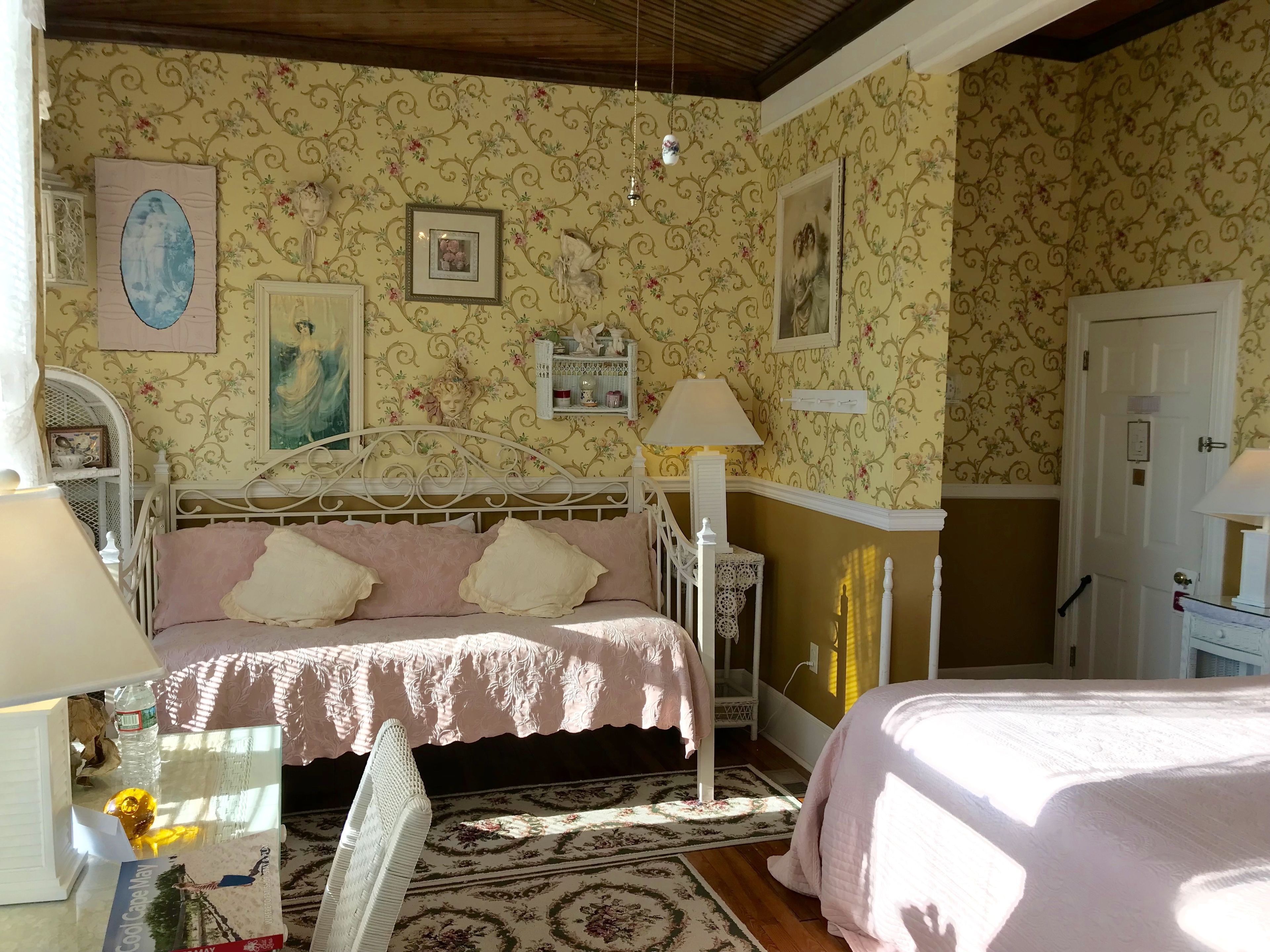 Ein Schlafzimmer mit floraler Tapete, einem Bett mit rosa Bettdecke, einem Nachttisch mit Lampe und einer Kommode.