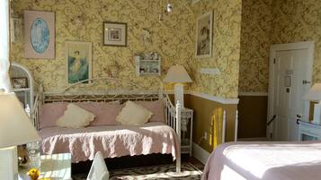 Ein Schlafzimmer mit floraler Tapete, einem Bett mit rosa Bettdecke, einem Nachttisch mit Lampe und einer Kommode.