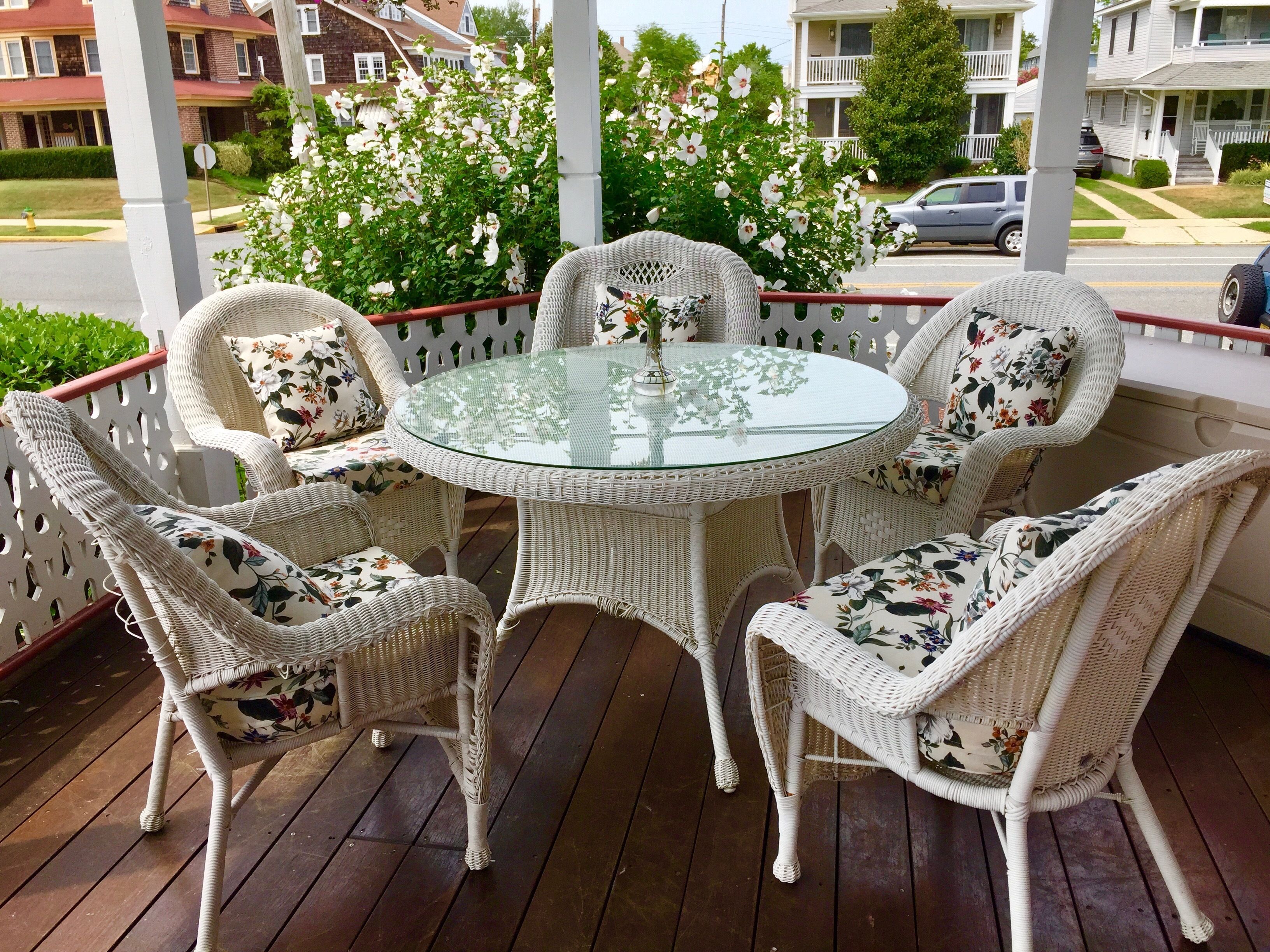 Ein runder Glastisch, umgeben von Korbstühlen mit floralen Kissen auf einer Holzterrasse.