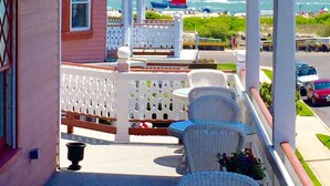 A porch with white wicker chairs and a small table, overlooking a beach with boats and people.
