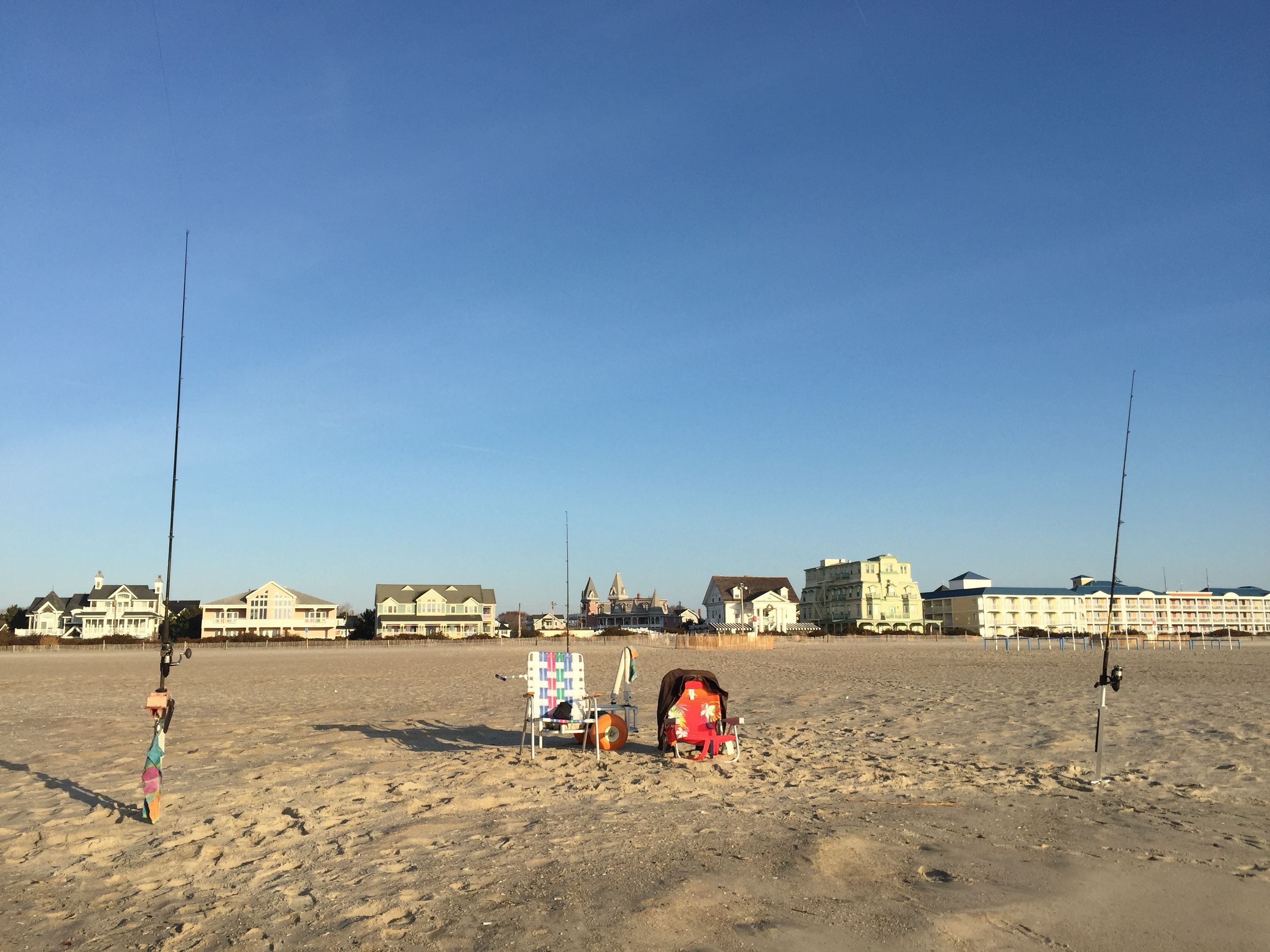 Una playa con dos cañas de pescar, una silla y un enfriador. En el fondo, varios edificios, incluido un hotel grande de varios pisos.
