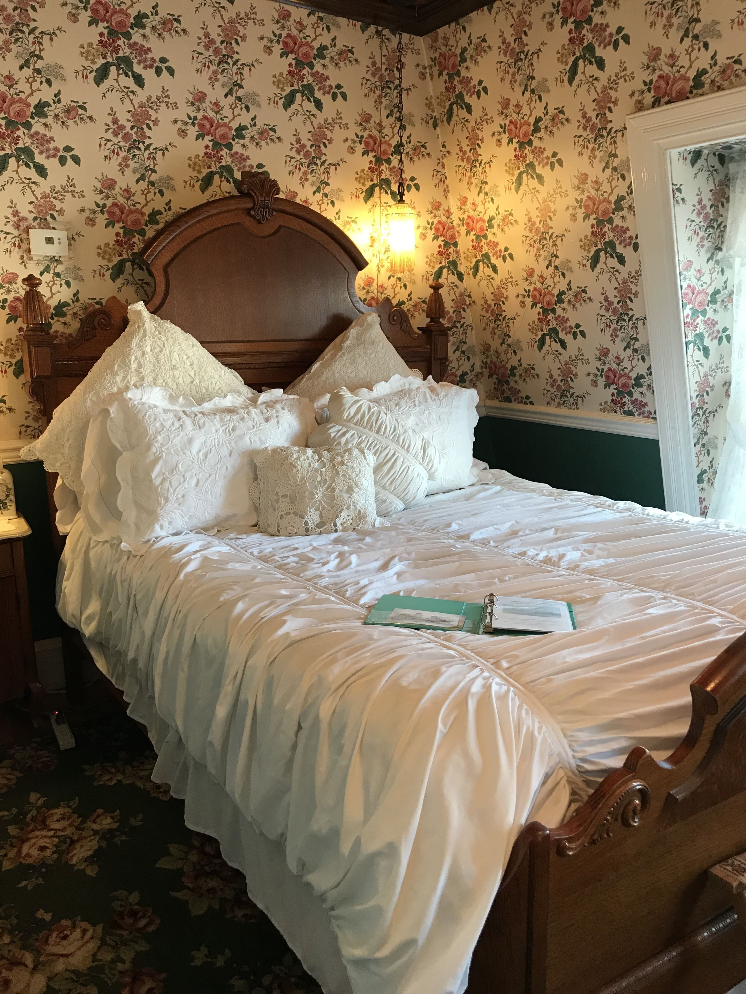 A neatly made bed with white linens and lace pillows, a wooden headboard, and a green book on the bed.