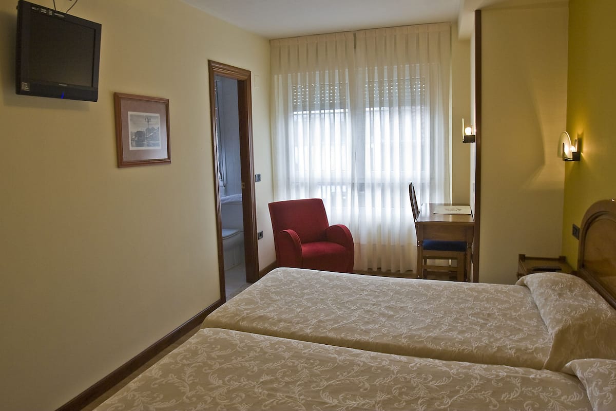 Habitación de hotel con dos camas, un televisor, un cuadro enmarco, una silla roja, un escritorio y una ventana con cortinas.