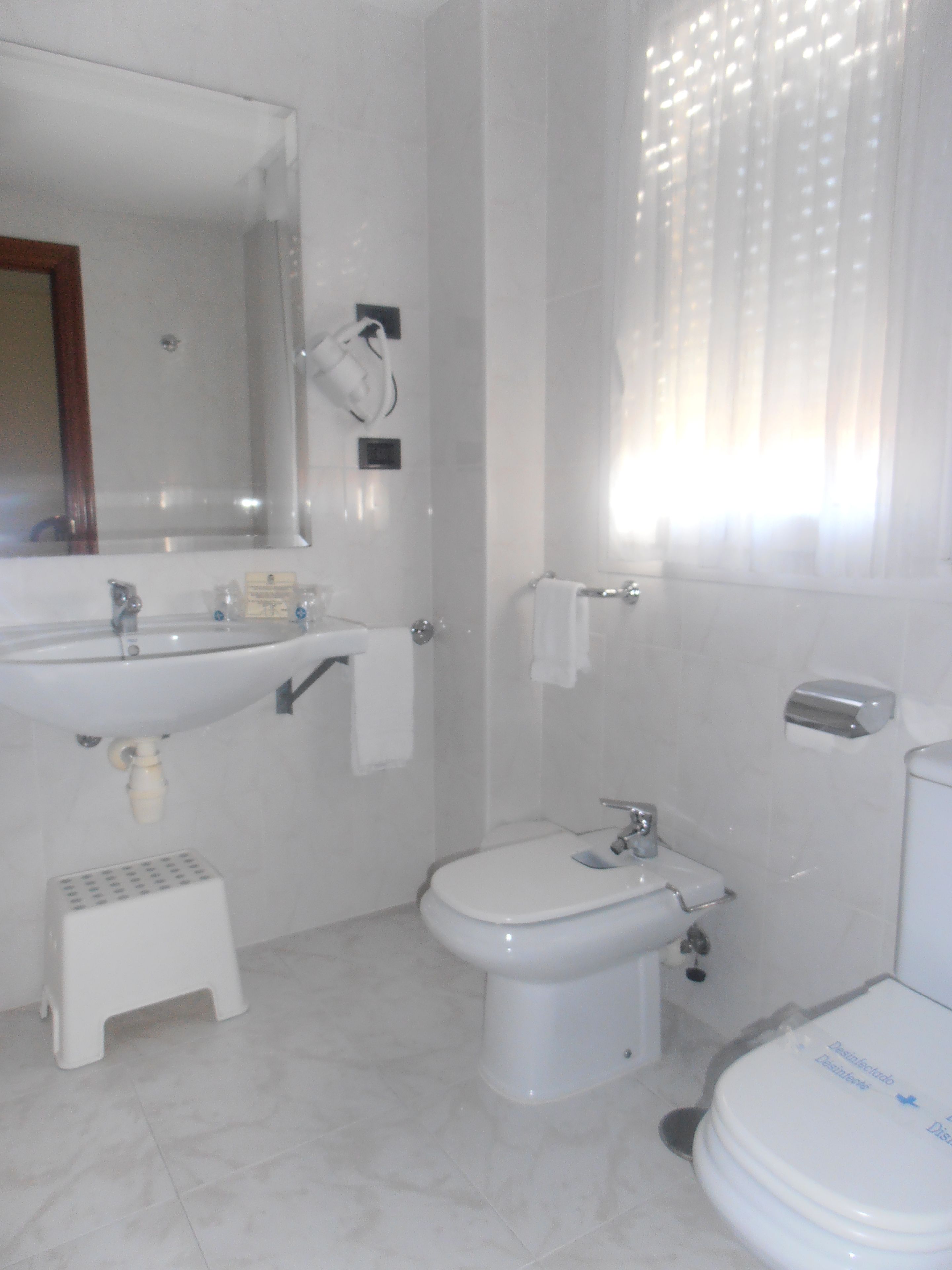 Un baño con lavamanos, inodoro y bidé. Hay un taburete cerca del lavamanos y un perchero en la pared.