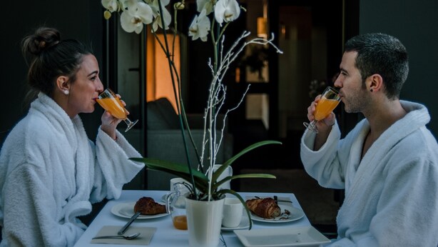 A couple in white robes sitting at a table with a white tablecloth, enjoying orange juice. There are pastries and a white pitcher on the table. - Ellauri Hotel, Landscape SPA - Adults Only (Zeanuri)