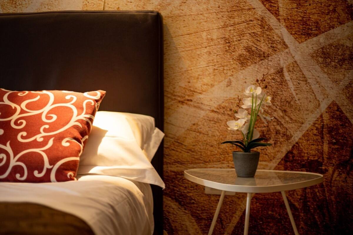 A neatly made bed with a patterned pillow, a small round table with a potted orchid, and a textured wall in the background.
