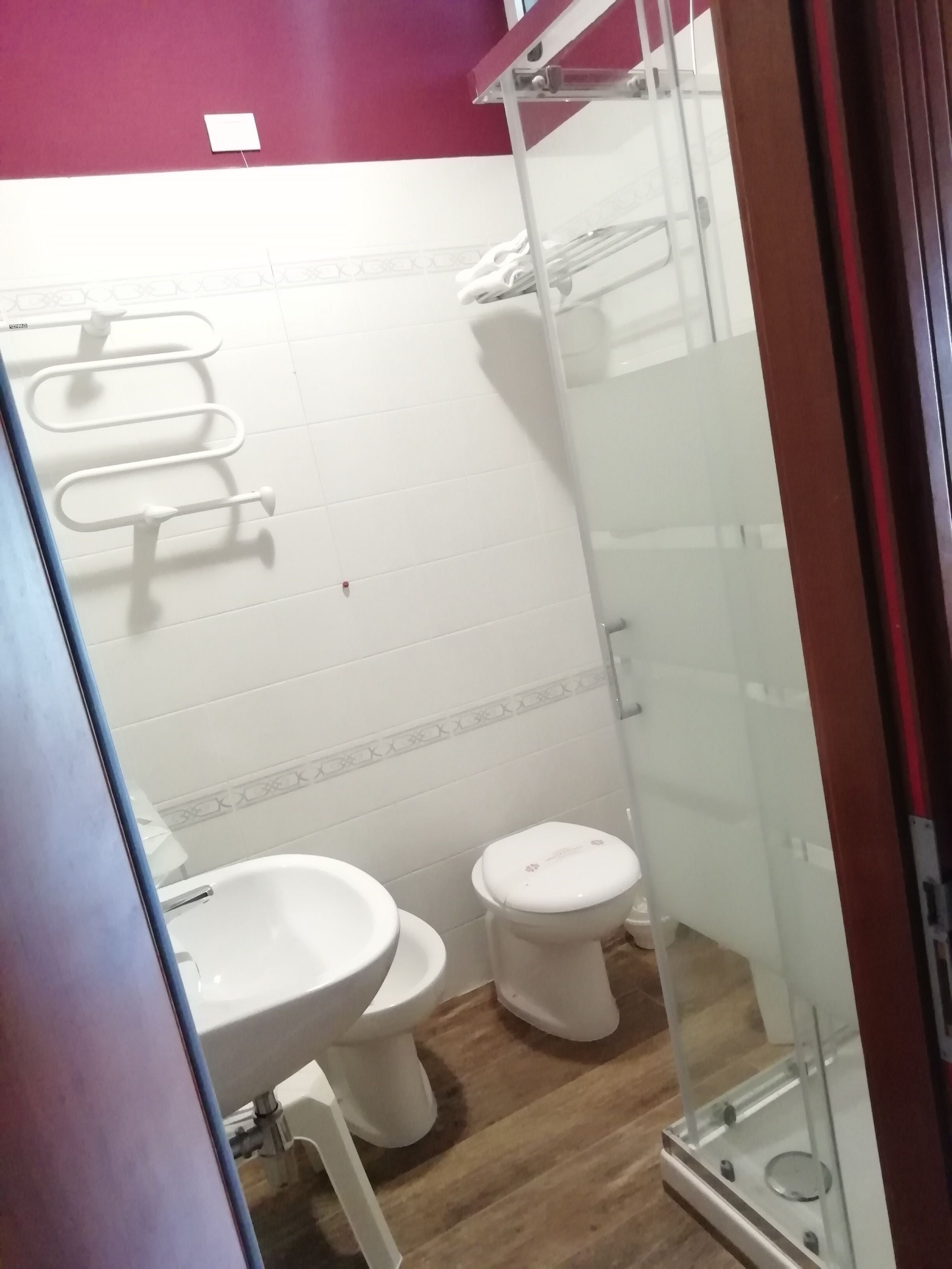 A bathroom with a white sink, toilet, and shower. There is a towel rack above the sink and a small shelf with toiletries.