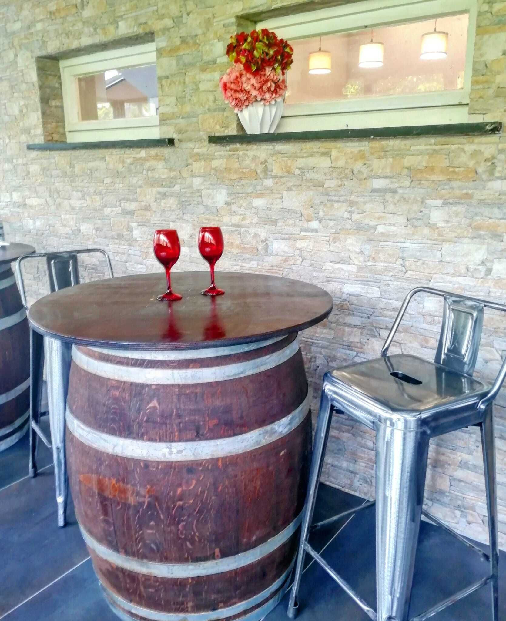 A round table with two red wine glasses and a large wooden barrel. There are metal chairs around the table, and a stone wall with a window above.