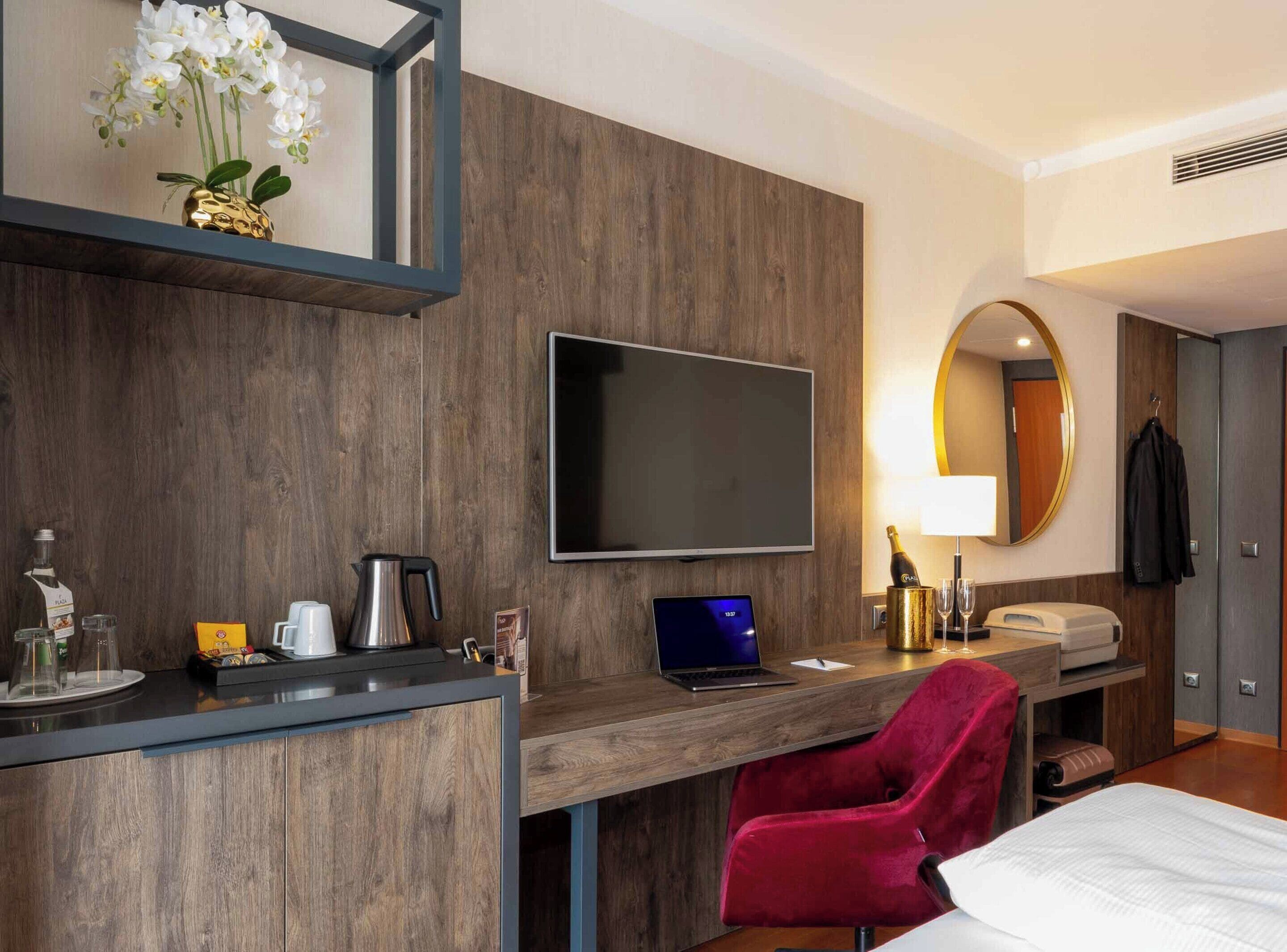 Habitación de hotel con escritorio de madera, una computadora portátil, una silla roja, un televisor, un minibar y una cama.