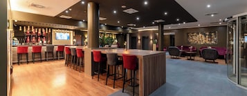 Zona de bar moderna con mostrador de madera, taburetes rojos y grises, y una pantalla grande en la pared.