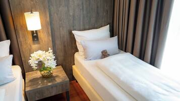 A hotel room with a wooden headboard, a bedside table with a lamp, a vase of white flowers, a bed with white bedding, and a teddy bear.