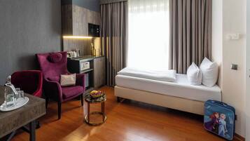 A hotel room with a sofa bed, a desk with a chair, a small table with a fruit bowl, a minibar, and a suitcase.