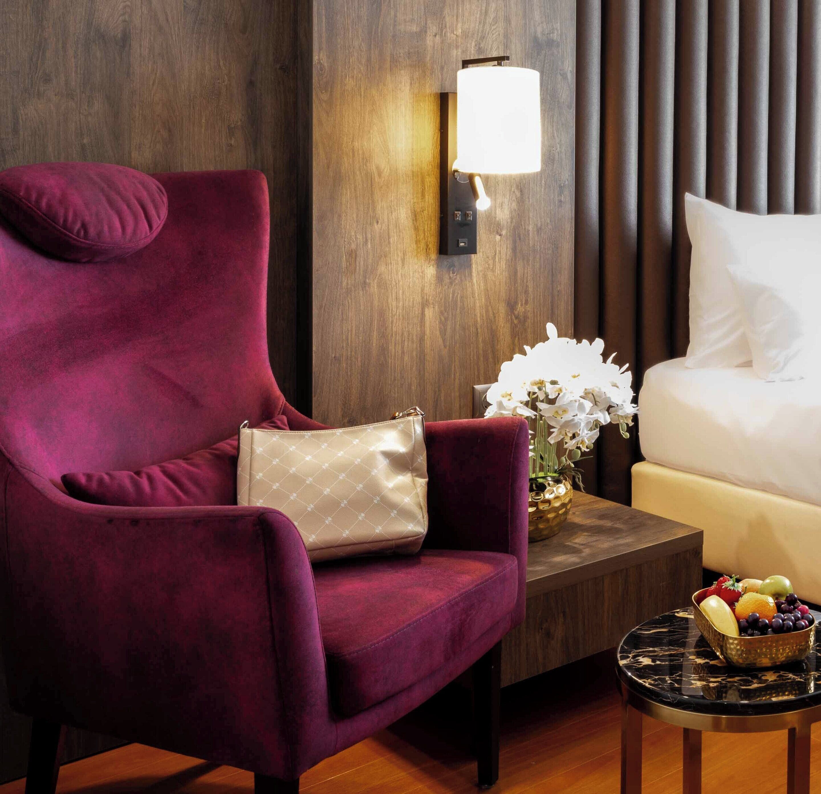 A plush red armchair with a beige handbag, a white sofa, a wall-mounted lamp, a small table with a fruit bowl, and a vase with white flowers.