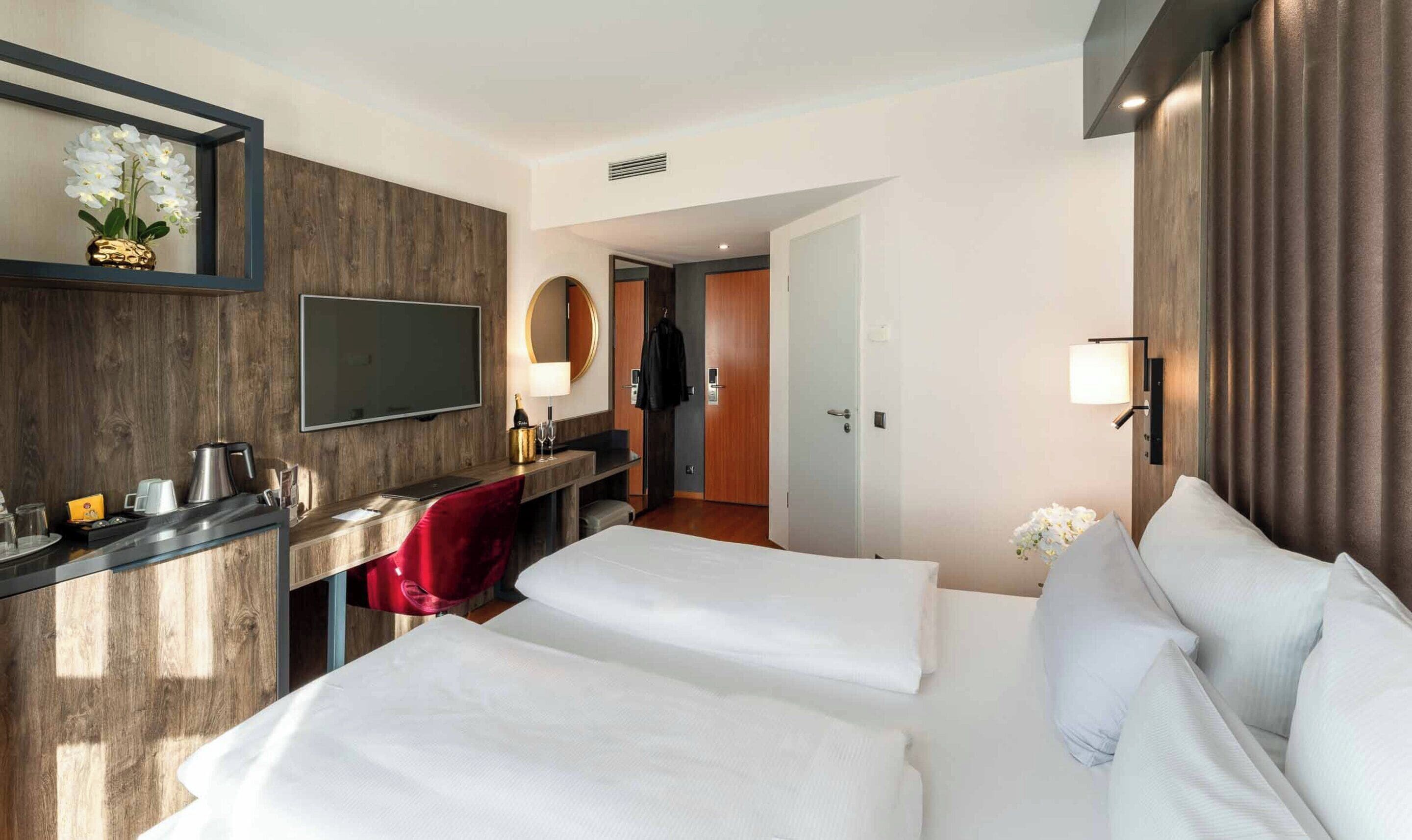 A hotel room with two beds, a desk with a flat-screen TV, a minibar, a mirror, and a door leading to a bathroom.