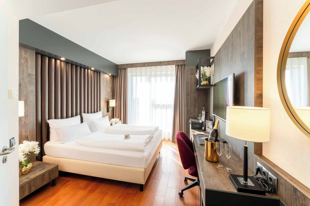 A modern hotel room with a large bed, a desk with a computer, a chair, and a minibar.