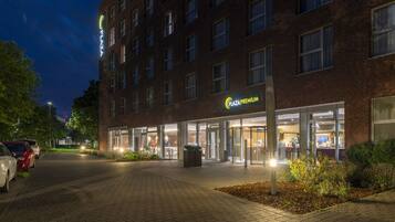 Ein Hotel Eingang mit einem 'Plaza Premium' Schild, einer Ziegel-Fassade und einem Parkplatz mit Autos.