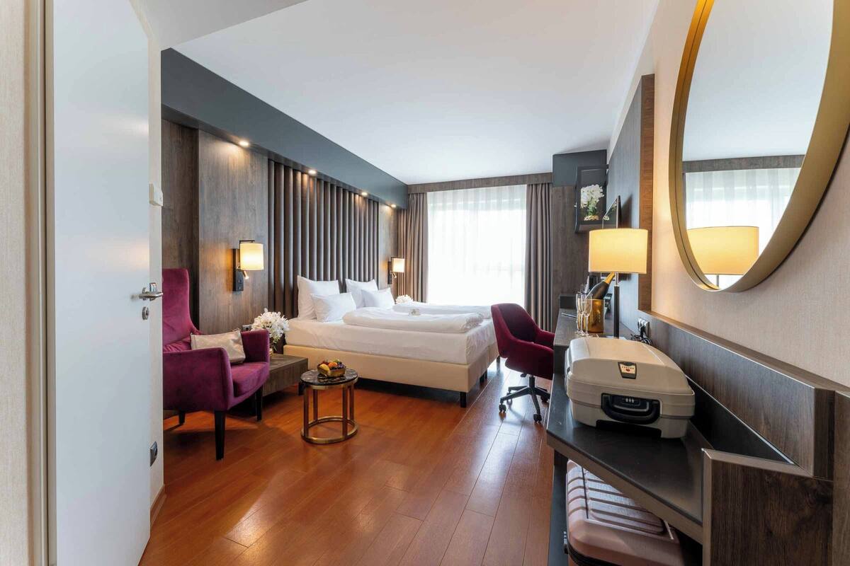 A hotel room with a double bed, a desk with a printer, a chair, a small table with flowers, and a mirror on the wall.