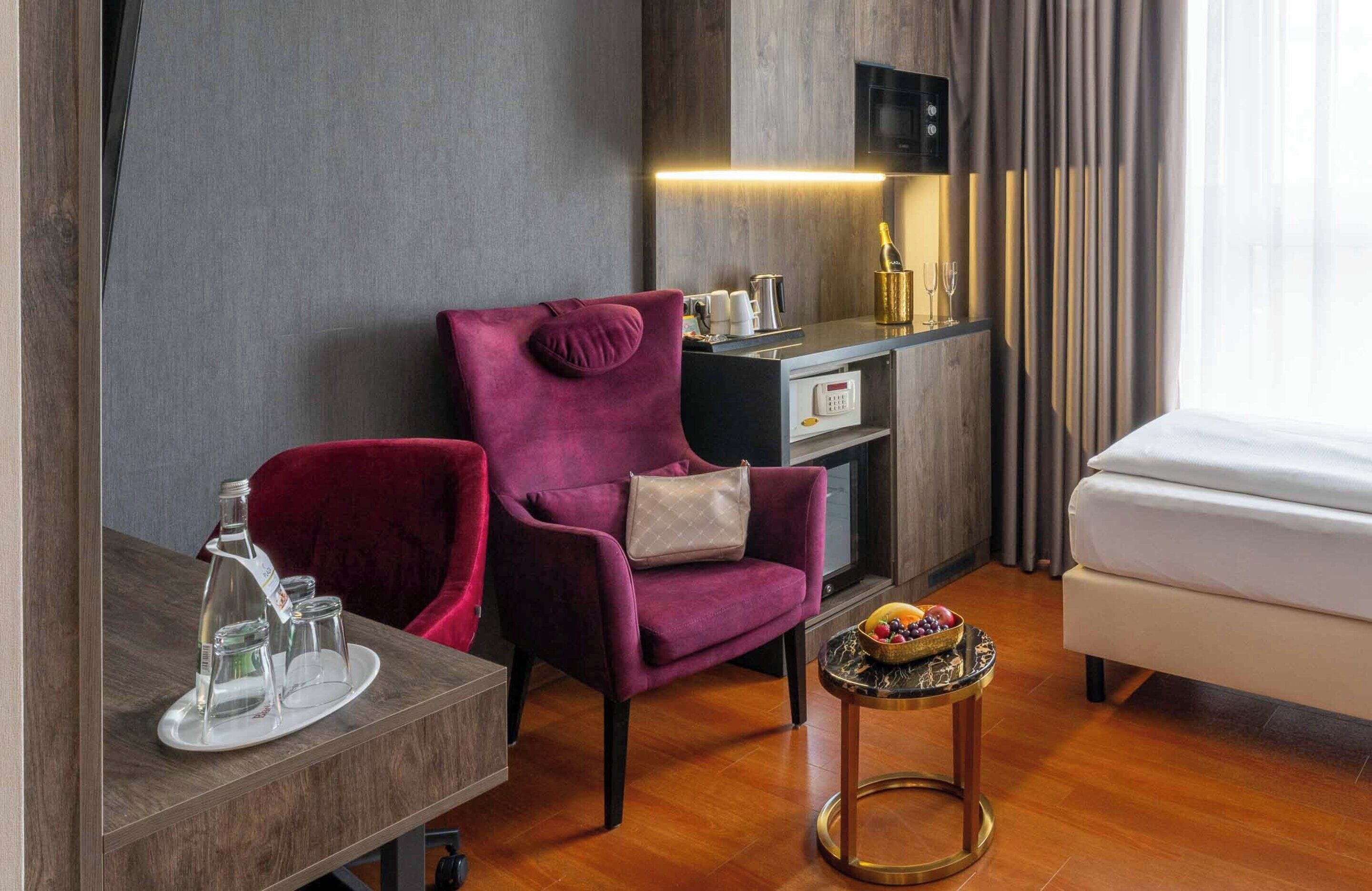 Una habitación de hotel con piso de madera, un sillón morado, una mesita redonda con fruteros y una minibar, además de una cama.