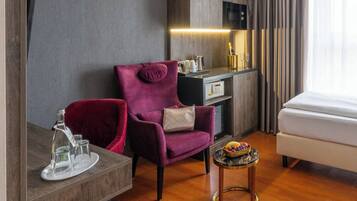 Una habitación de hotel con piso de madera, un sillón morado, una mesita redonda con fruteros y una minibar, además de una cama.