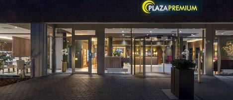 The exterior of Plaza Premium with a glass entrance, indoor seating visible through the doors, and a well-lit facade.