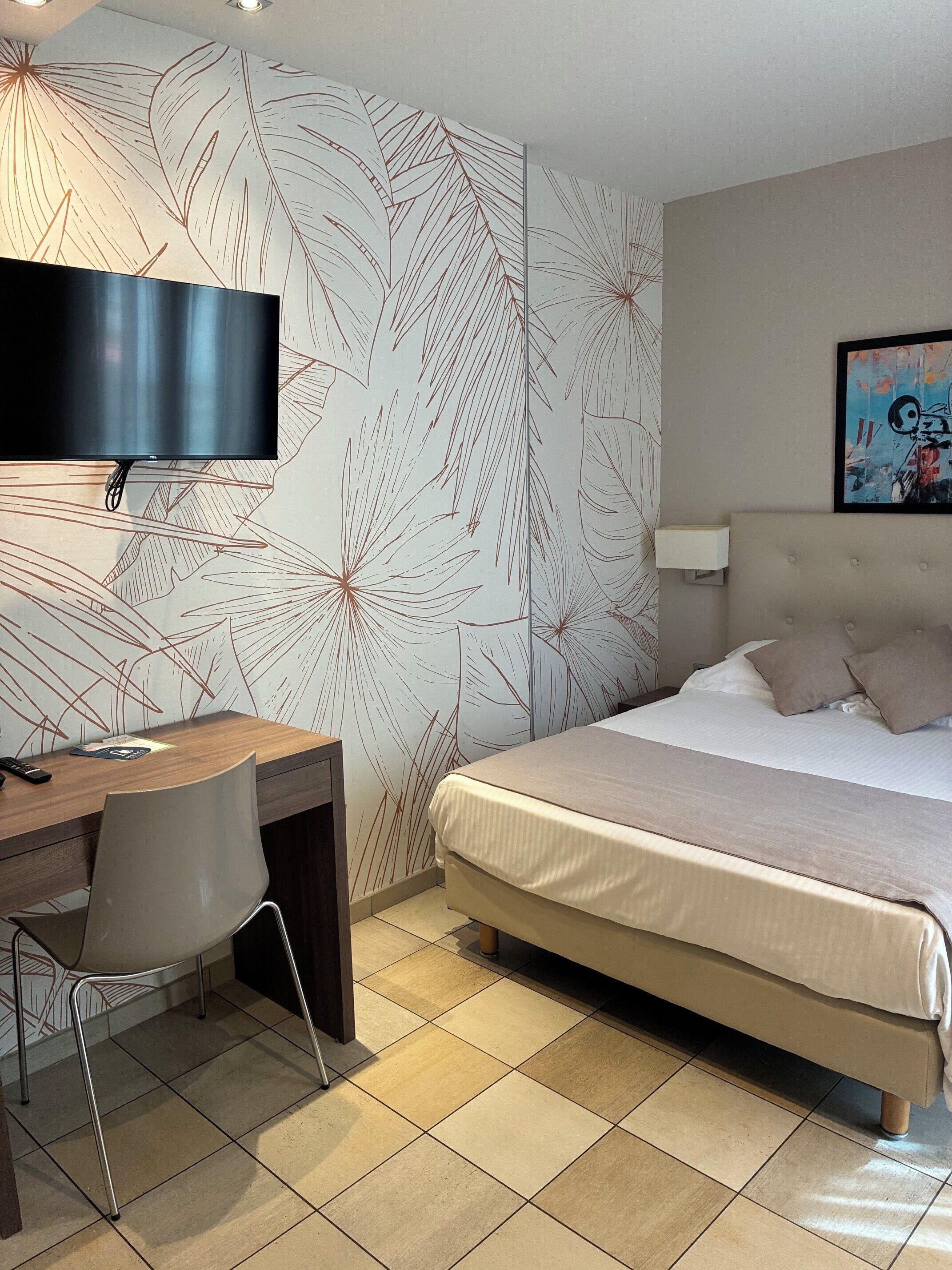 A hotel room with a bed, a desk with a chair, a television, and a wall with a leaf pattern.