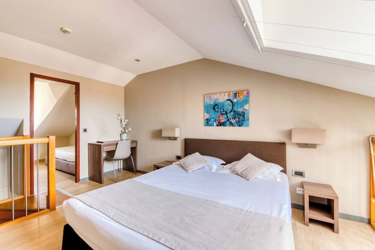 A bedroom with a large bed, a desk, a chair, a painting on the wall, and a small table.