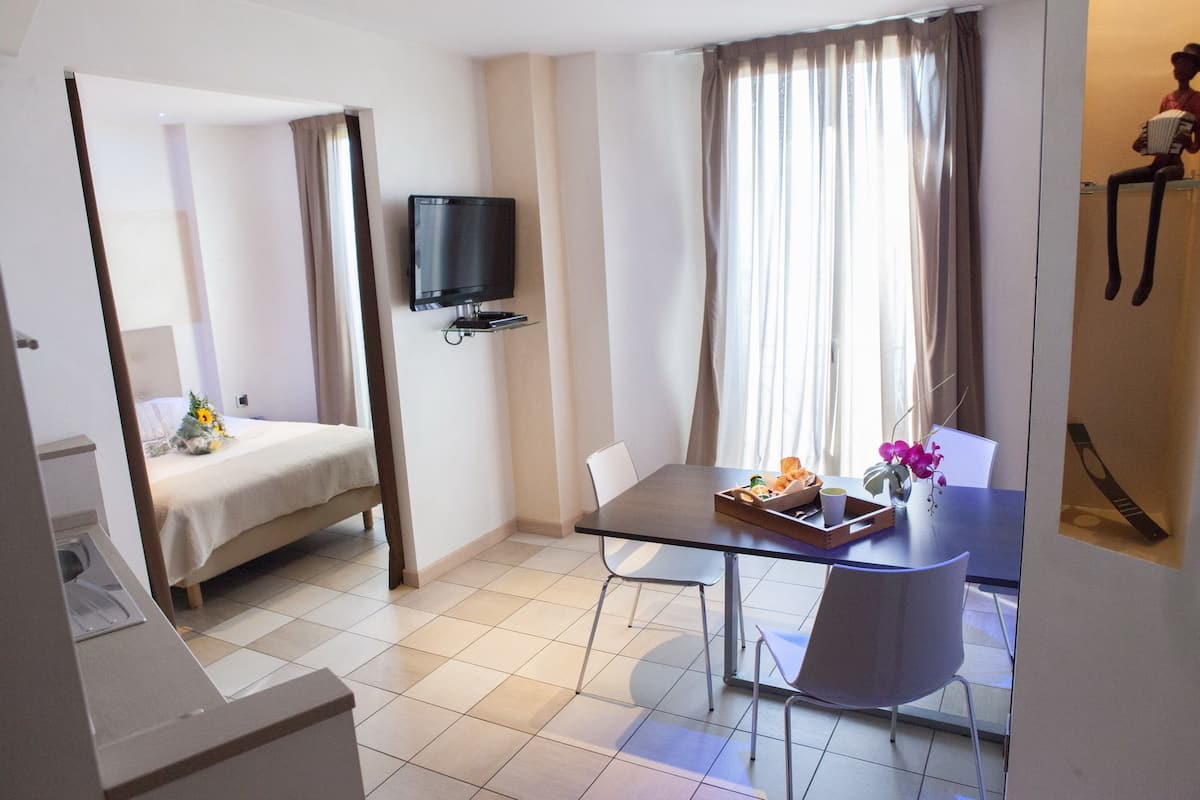 A hotel room with a bed, a TV mounted on the wall, a dining table with chairs, and a window with curtains.