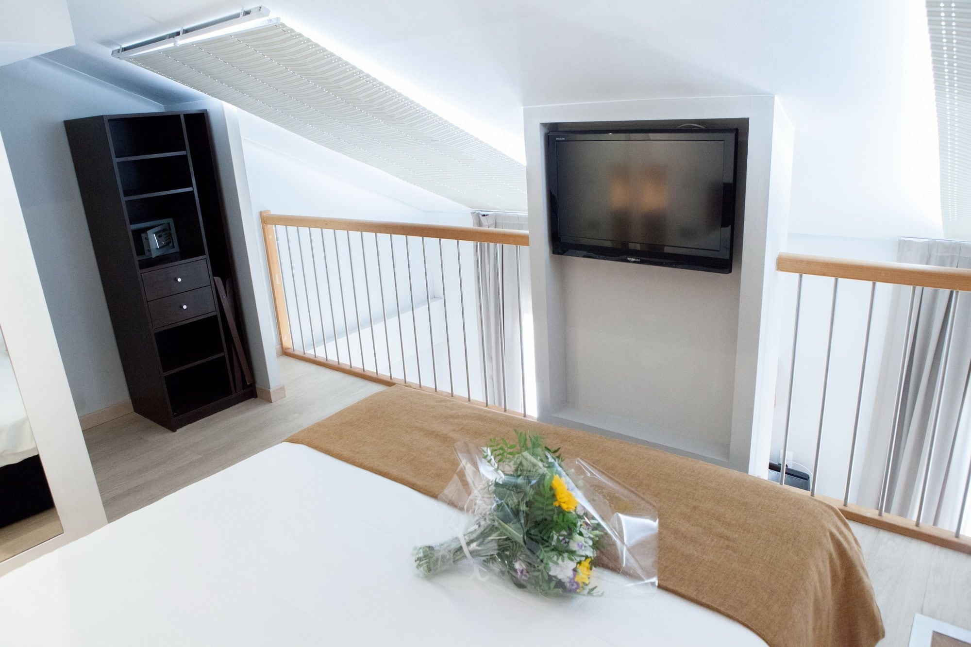 A modern hotel room with a bed, a TV mounted on the wall, a wooden wardrobe, and a balcony with a white railing.