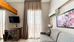 A modern living room with a grey sofa, a wooden desk, a TV, and a large window with curtains. - Hotel La Villa d'Elsa (Antibes)