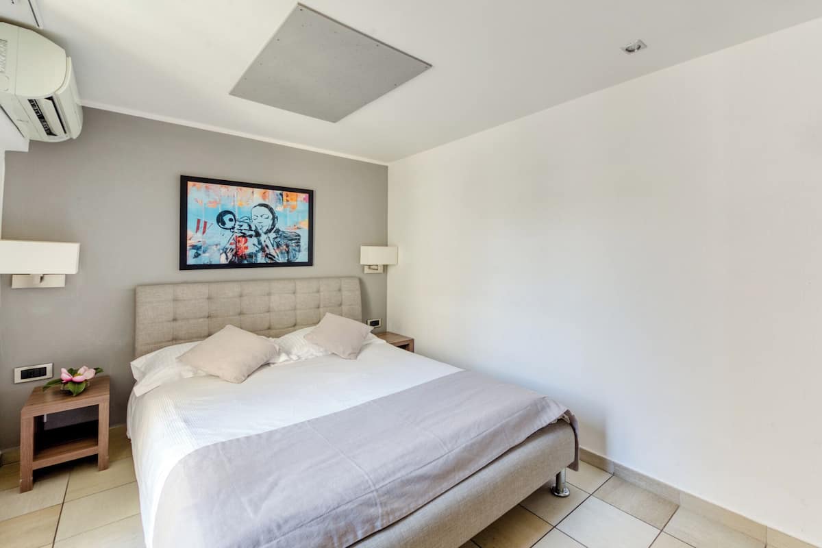 A modern hotel room with a large bed, two bedside tables, a wall-mounted air conditioner, and a ceiling light fixture.
